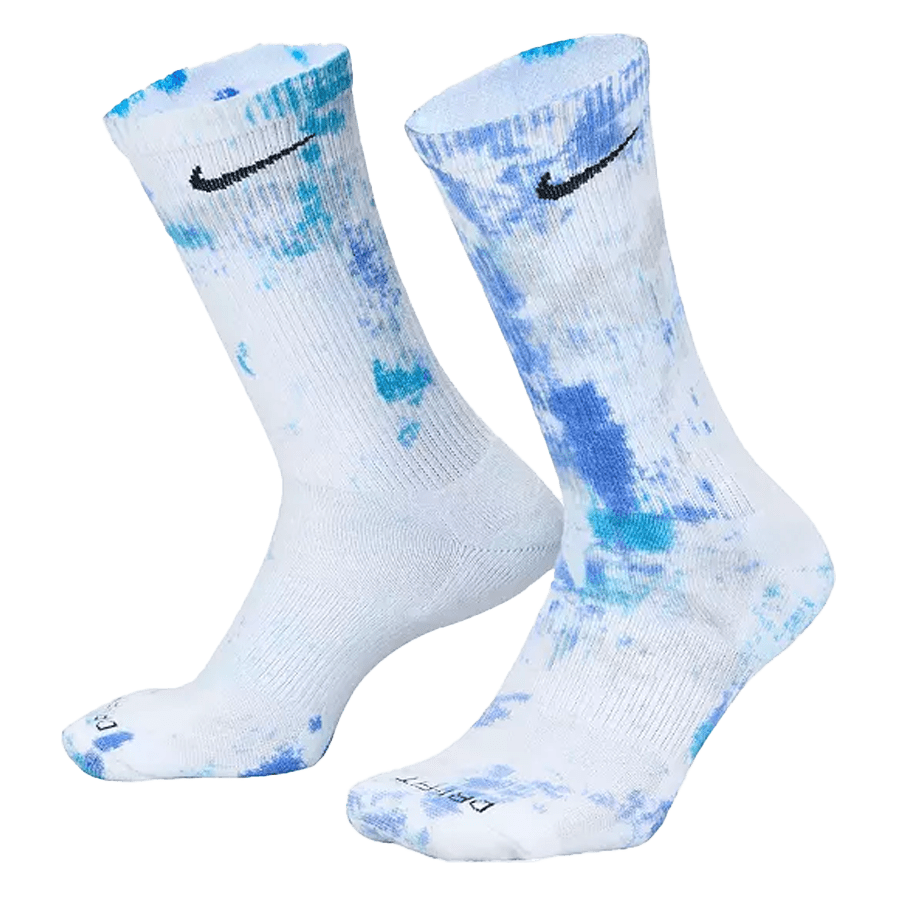 The Nike Everyday Plus Tie-Dye Socks, designed in white with blue tie-dye patterns and featuring a black Nike logo near the top, are displayed standing upright to highlight their design and length.